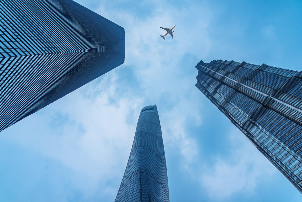 A plane flying in the sky with buildings in the forefront.